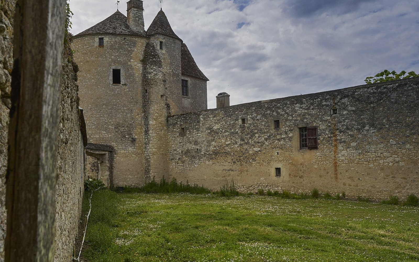 Stone turrets and fortified wall of a château rise above an overgrown grassy courtyard